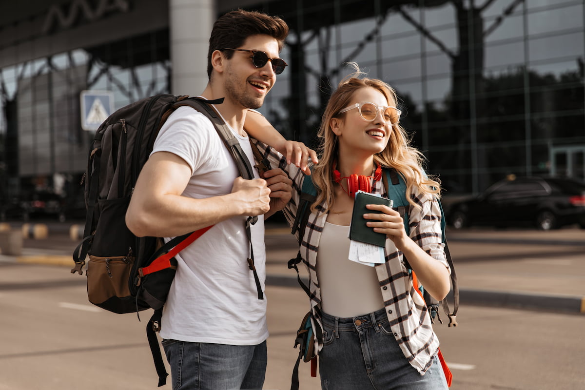 Travelers with backpacks poses near modern airport. Happy blonde woman in plaid shirt, sunglasses h